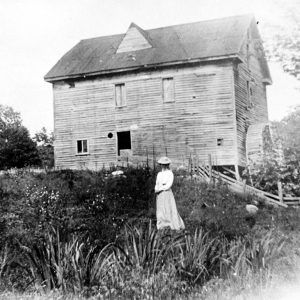 young woman in dress and hat standing before multistory wooden building