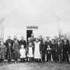 Group of white children and teachers standing in front of wooden school building