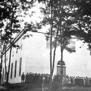 Multistory wooden building with large trees and a group of people in front