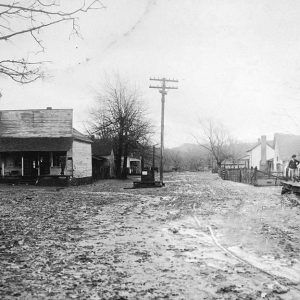 muddy dirt street with wooden buildings and some people standing on a porch