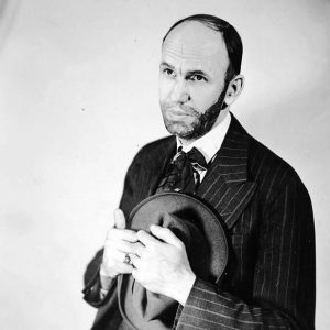 Man in dark pinstripe suit crushes hat to chest