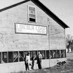 Two white men standing before a large wooden building "Lincoln Co-op Fruit Vegetables"