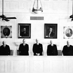 Seven formally dressed white men standing in front of a long judicial bench with framed portraits behind them