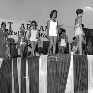 White man at microphone and white beauty pageant contestants in bathing suits with numbers pinned to them