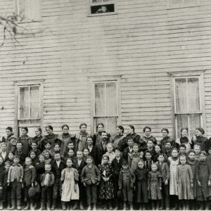 Large group of white children posing with teachers in front of multistory building with white exterior and child looking out through top window