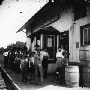 adults and children standing on the deck of a train depot "Cave Springs" with barrels stacked nearby