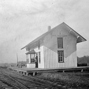 man standing on porch of small wooden train depot building