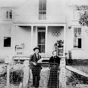 Elderly white couple standing before a multistory house with another white woman standing inside looking out