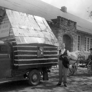 White man standing by tiny school replica mounted on back of truck with sign on it saying "the school of yesterday"