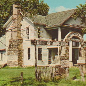 Multistory white house with stone chimney and fronted by columns with sign "Pea Ridge Battlefield" mounted before it