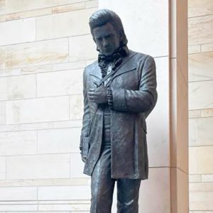 Bronze statue of man wearing a suit with a tuxedo shirt and bow tie. He has a guitar strap around him and is holding his guitar to his side