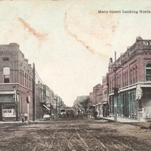 Postcard featuring a dirt street running between two-story business buildings "main street looking north"