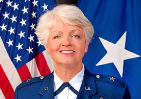 White woman with white hair dressed in blue military uniform in front of American flag and Air Force Brigadier General flag