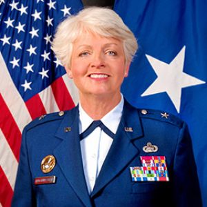 White woman with white hair dressed in blue military uniform in front of American flag and Air Force Brigadier General flag