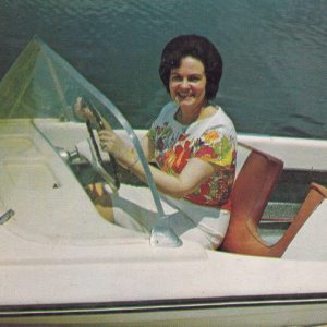 White woman seated smiling while holding steering wheel in speed boat on a lake