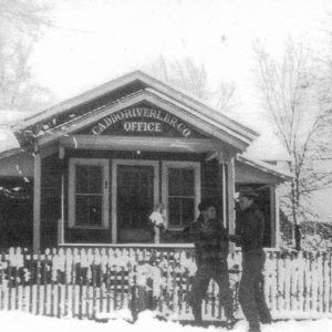 White man holding a snowball up to another white man standing in front of single story building behind wooden fence covered in snow