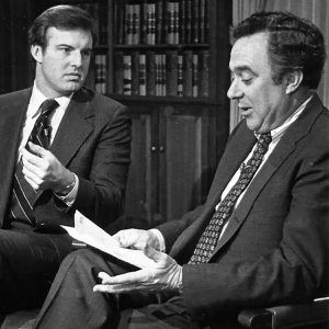 Two white men in suits conversing while one looks down at document in hands