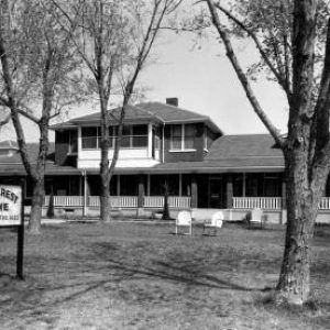 Multistory residential house with large yard and several trees two chairs and sign