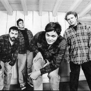 Four white men in flannel shirts posed in front of blank wooden wall between two windows