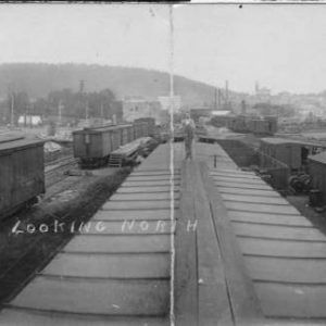 White man standing atop railroad car in middle of outdoor area containing railroad tracks freight cart carrying steel and a small building with city view in background inscription "looking north"