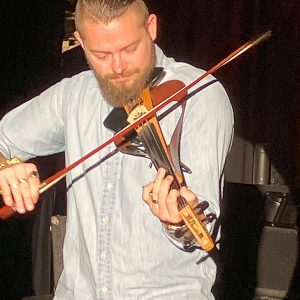 White man playing a violin onstage under a spotlight