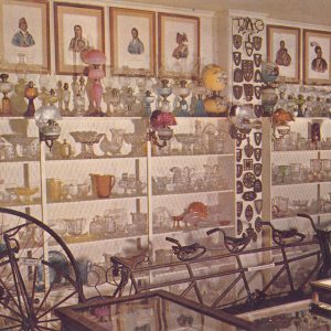 Wide variation of glassware on shelves behind various old bicycles oil lamps metal decor and framed portraits on display