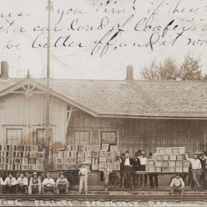 White men and a few boys in front of a singe-story shipping building and carts with piles of boxes