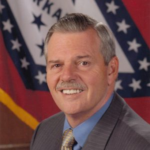 White man with mustache wearing suit and Arkansas pin in front of state flag