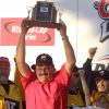 White man with mustache wearing glasses standing in front of small cheering crowd holding trophy above his head smiling