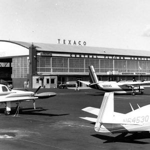 Several airplanes parked near large aircraft garage building with sign "Texaco"