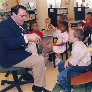 White man sitting holding open book reading to multiracial group of children at nearby tables in children's section of library