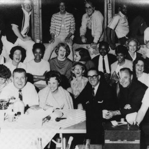 Large group of black and white men and women smiling behind beverages small ice buckets and telephone set on table
