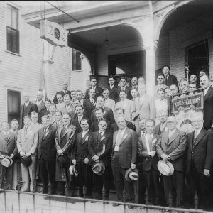 Group of white men standing along the steps of a multi story brick building in four rows