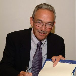 Older white man with mustache and glasses wearing suit holding sharpie and open book with signature signed inside
