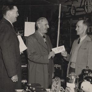 Two older white men in dress coats one handing pamphlet titled "Brief Facts about Rotary" to white man in sweater and jacket inside store