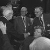 Older white man back turned shaking the hand of another white man standing next to a third white man wearing medal while older white woman seated looks up smiling