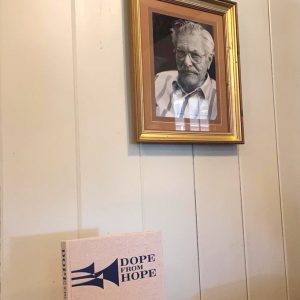 Bound book on table underneath framed photo of older white man with mustache wearing glasses
