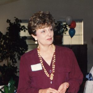 White woman wearing name tag and long beaded necklace speaking to older white man in suit and glasses