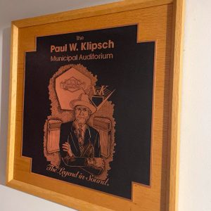 Framed wooden plaque with portrait of older white man wearing glasses suit and hat standing arms crossed in front of two speakers above text "the legend in sound"