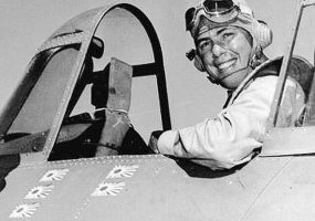 White man wearing flight helmet sitting in aircraft cockpit smiling