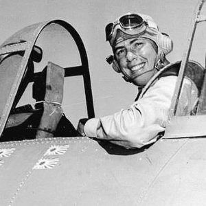 White man wearing flight helmet sitting in aircraft cockpit smiling