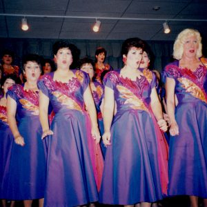 Group of white women wearing matching decorative dresses and dangling earrings singing