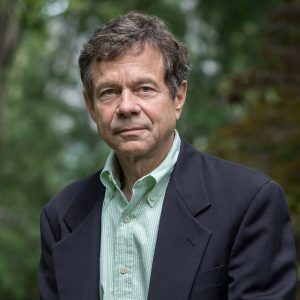 Middle aged white man in striped button up shirt and suit jacket in front of blurred forest background