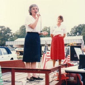 Middle aged white woman standing on flatbed trailer with rails speaking into microphone another woman standing between two American flags smiling behind her
