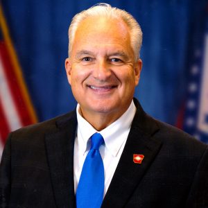 Older white man clean shaven in suit and tie wearing Arkansas state pin smiling