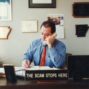 White man in dress shirt and tie holding rotary phone to ear in office sitting behind desk with sign "The scam stops here"