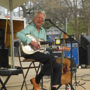 Older white man wearing button up shirt slacks and dress shoes sitting in metal chair on stage surrounded by sound equipment playing metal guitar singing into microphone