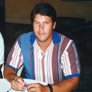 White man dressed in vertical stripped collar shirt sitting at table holding pen