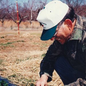 White man in sunglasses camouflage jacket and hat crouched down inspecting plant