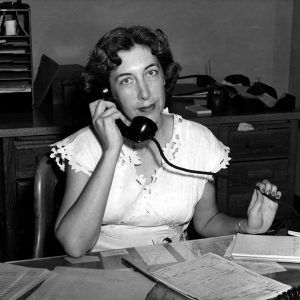 White woman short curled hairstyle sat at desk answering rotary phone